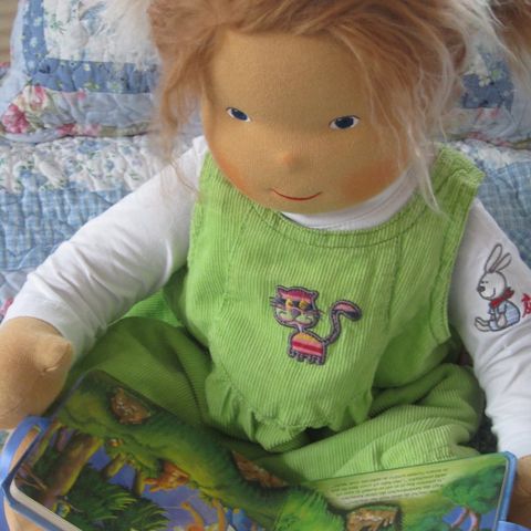 Große Babypuppe mit rotblondem Haar in grüner Latzhose, ein Bilderbuch auf dem Schoß