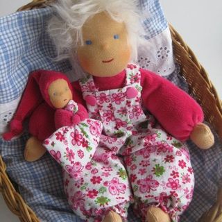 Weißblonde Stoffpuppe in geblümter Latzhose in einem Weidenkorb, daneben ein rotes Püppchen
