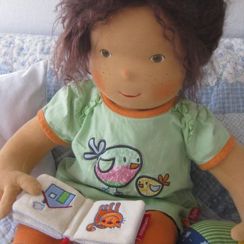 Babypuppe mit dunkelbraunem Haar in grünem T-Shirt mit einem Stoffbuch