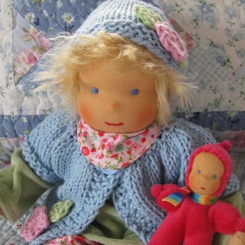 Blonde Gliederpuppe mit gestrickter Mütze und hellblauer Strickjacke, daneben ein rotes Püppchen
