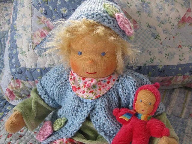 Blonde Gliederpuppe mit gestrickter Mütze und hellblauer Strickjacke, daneben ein rotes Püppchen