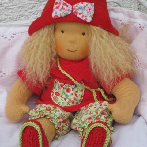 Blonde Gliederpuppe mit rotem Hut, geblümter Hose und gehäkelten roten Schuhen, sitzend
